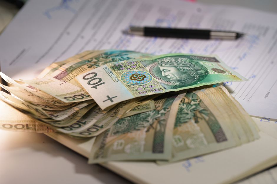 A close-up view of several British pound banknotes, including £100, £50, and £20 denominations, placed on top of a stack of paper documents, with a black pen resting on the papers. The documents ap